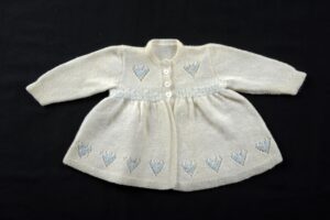Cream knitted childs matinee jacket with blue heart motifs on the chest and around the bottom on a cream stocking stich ground.