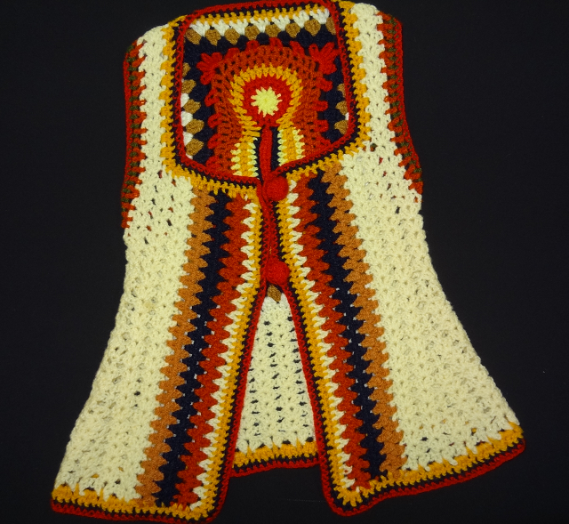 Crochet waistcoat with square neck, two large red buttons striped borders and thicker stripes either side of the opening. The back is visible through the neck opening to show that the central striped extend to just below the neck where they turn to form a closed box with a bright circle in the middle.