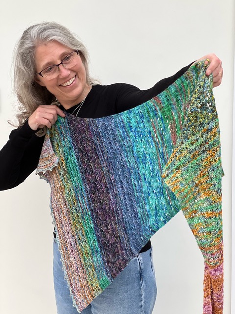 Emma Vining holding up her openwork scarf with bands of colour: Merrow Berries Shawl.