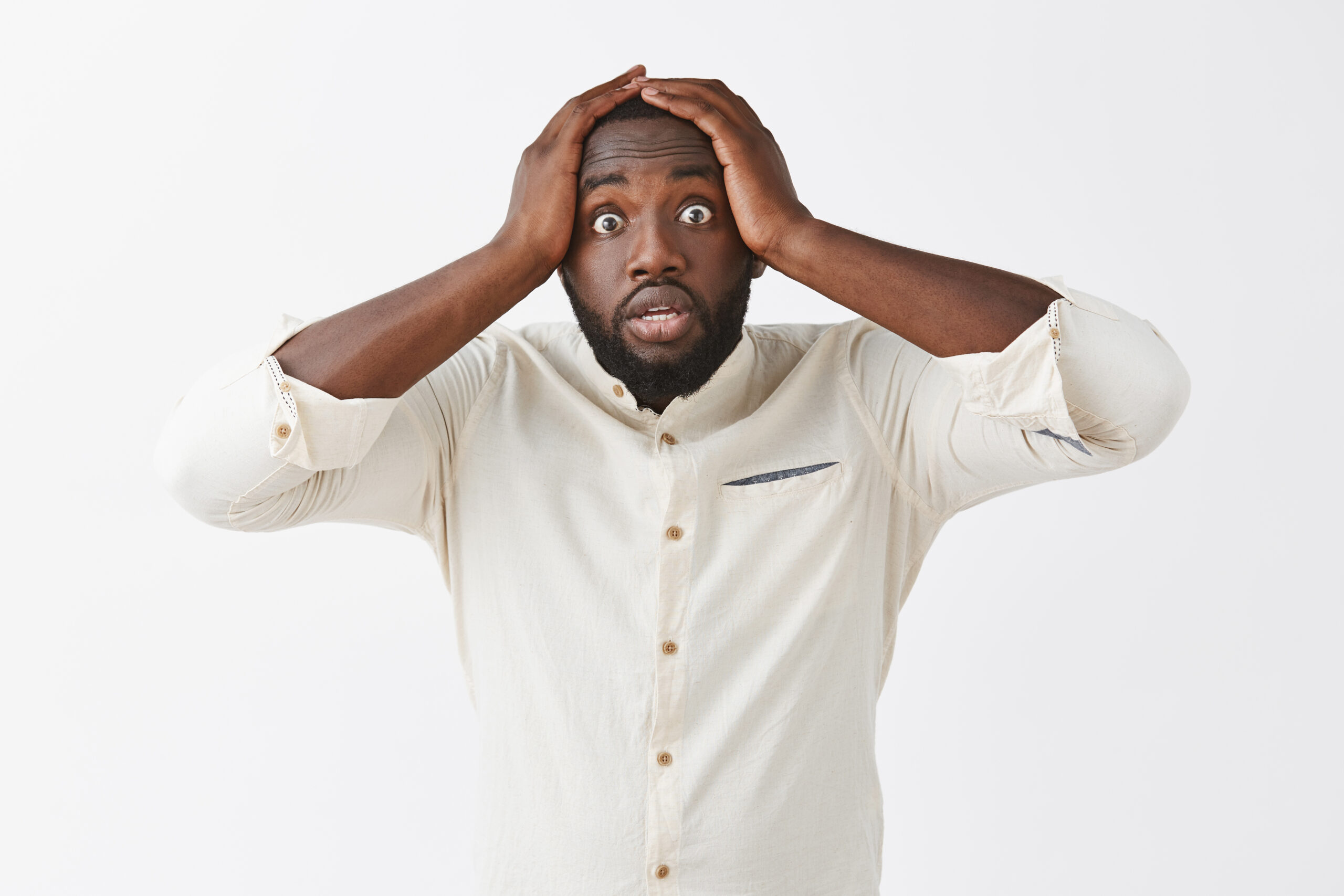 Portrait of worried cute dark-skinned male panicking, holding hands on head and starring with popped eyes at camera, being anxious or nervous over gray background.