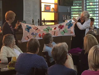 Two ladies showing crochet scarf with applique butterflies watched by fascinated crafters.