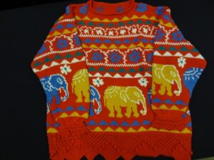 Red jumper with triangular welt with openwork and bobbles. Boey has stripes colourwork designs in blue, white, yellow and green on a red ground, the most dominant being a row of elephants.