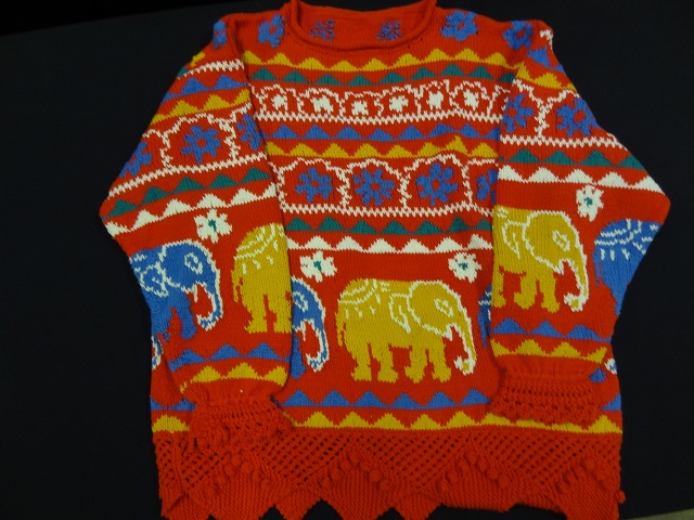 Red jumper with triangular welt with openwork and bobbles. Boey has stripes colourwork designs in blue, white, yellow and green on a red ground, the most dominant being a row of elephants.