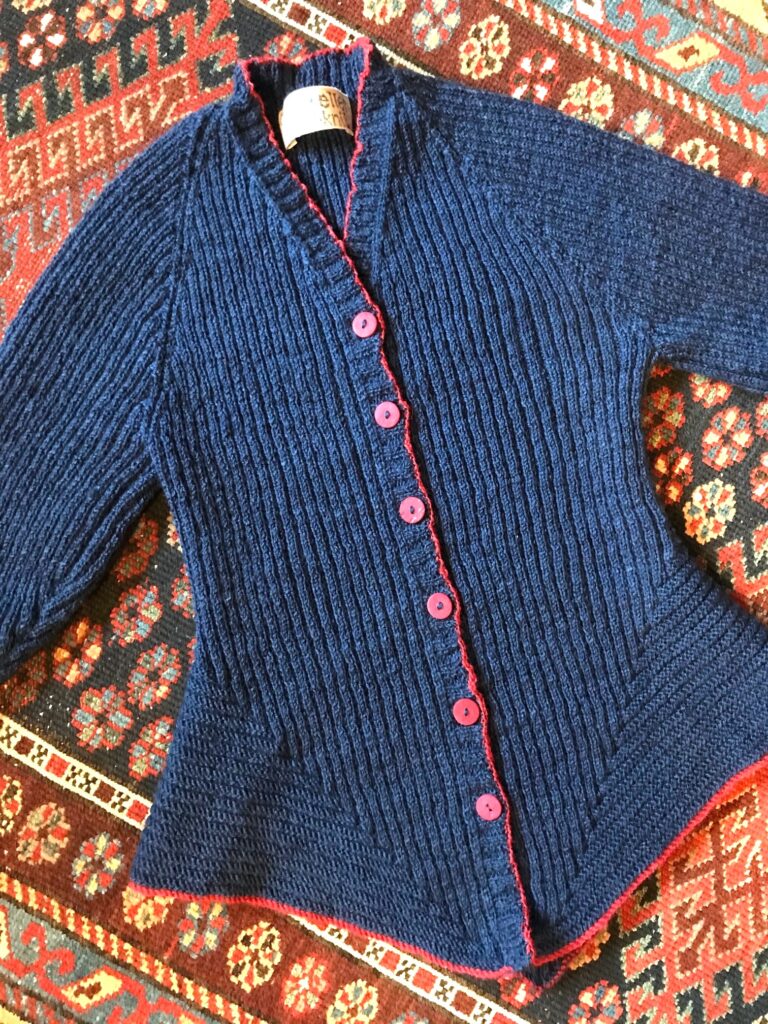 Blue cardigan with triangular bottom in blue with red edging. The body is in rib with a contrasting stitch pattern at the bottom.