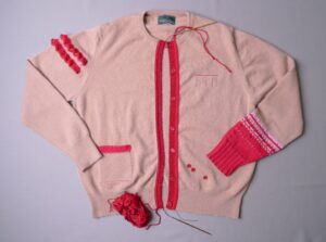 Photograph of a pink cardigan with dramatic red button bands, pocket top, cuff and arm decoration.