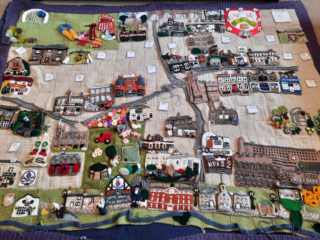 Photograph of the knitted Pudsey map under construction. Some landmarks have already been sewn into place, others are pinned into their planned positions, and written notes show where others are to be put.