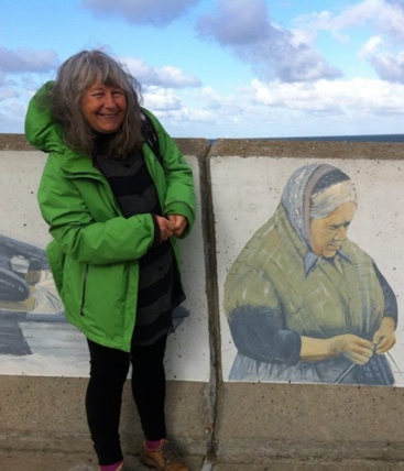 Rita Taylor standing against a wall with a painting of an old lady knitting