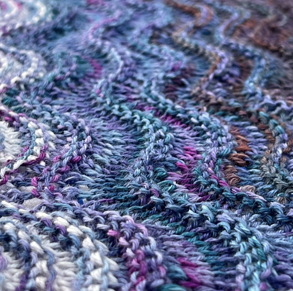 Knitted fabric in shades of blue and pink with garter ridges and waved created by increases and decreases.