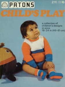 Toddler wearing knitted striped jumper, shorts and bootees. Sitting next to stuffed "Humpty" toy.