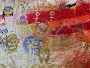 Photograph of a section of textile art with fabric dyed in broad stripes nd blocks with embroidered faces resembling sketches by children of varying ages.