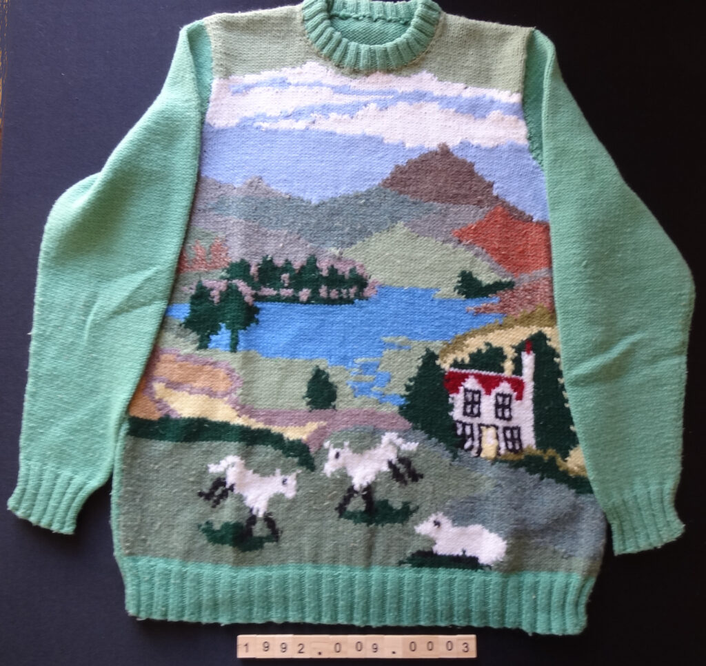Sweater with high round neck and set in sleeeves, welt, cuffs and double neck band in double rib. Front patterned with scene of hills, lake, house and sheep.