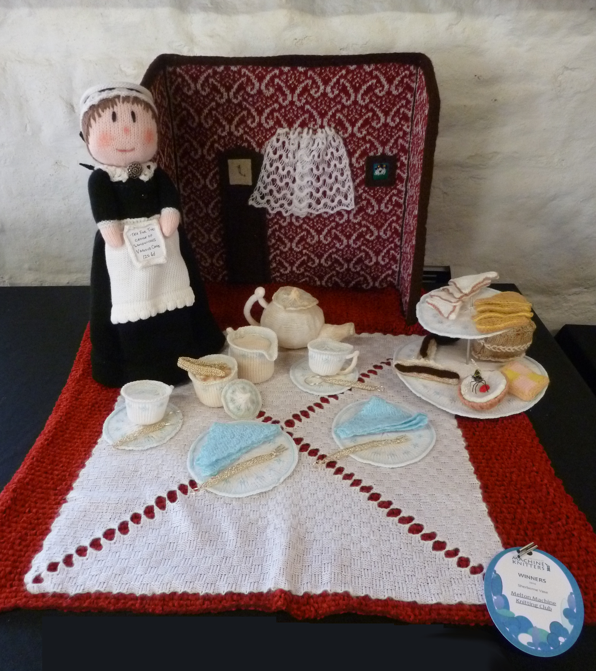 Machine knit scene of a traditional afternoon tea complete with carpet, wall and curtains, tablecoloth, waitress, cake stand, tea set, cutlery, napkins and, of course, sandwiches and cakes. A knitted fly is also enjoying the spread.