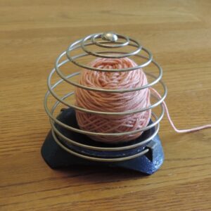 Photograph of a "Princess" yarn holder - a spiral of wire on a blue metal base - enclosing a ball of pink yarn.
