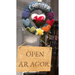 Knitted wreath with heart featuring Ewlsh dragon in centre from which hangs a wooden sign saying Open/Ar Agor.