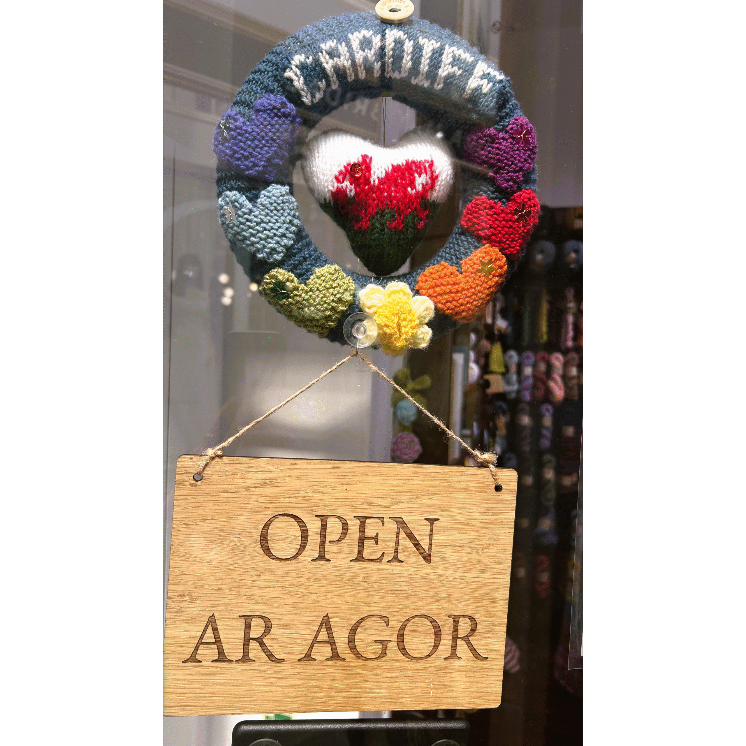 Knitted wreath with heart featuring Ewlsh dragon in centre from which hangs a wooden sign saying Open/Ar Agor.